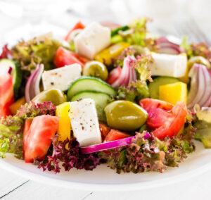 A hearty greek salad with cubes of vegan Feta cheese, tomatoes, cucumbers, olives and peppers on a bed or curly red lettuce