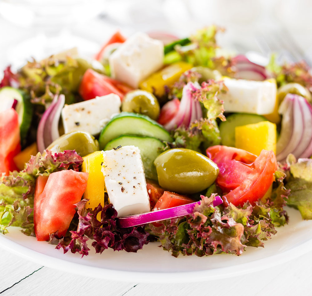 A hearty greek salad with cubes of vegan Feta cheese, tomatoes, cucumbers, olives and peppers on a bed or curly red lettuce