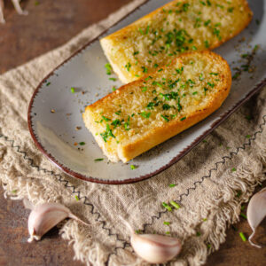 Broiled garlic bread, sprinkled with shaved vegan Parmesan on a stoneware plate with garlic cloves scattered arounf it